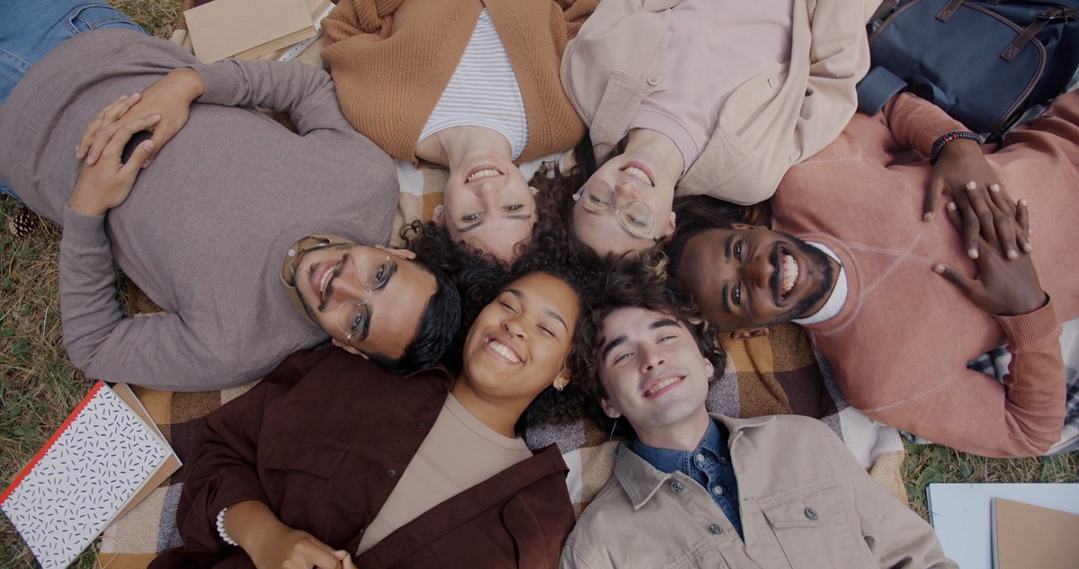 A diverse group of six friends is lying on a checkered picnic blanket in a circular formation, their heads touching in the center, discussing why some words are difficult to translate